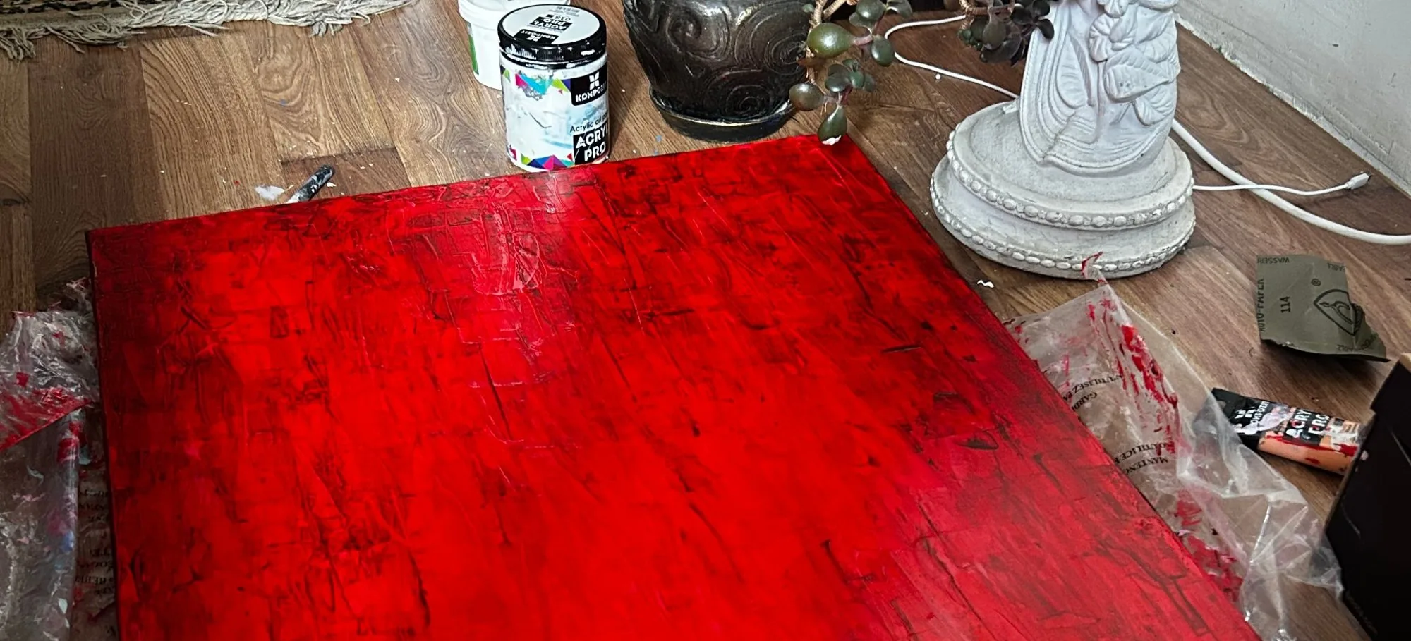 Crimson Dawn, a red textured abstract painting by Nataliia Ivaniv, lying on the studio floor beside paint jars and a potted plant